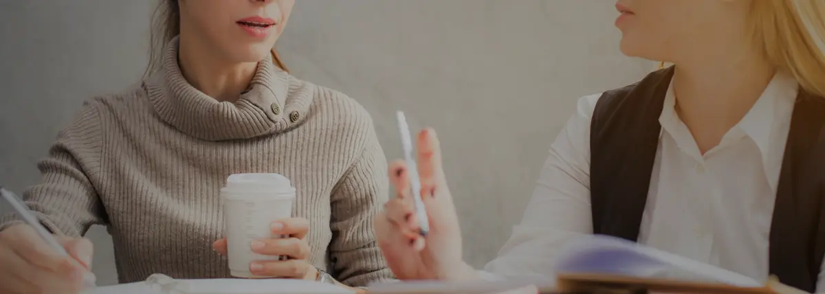 picture of two people sitting down at a table, one person holding a travel cup and the other holding a pen